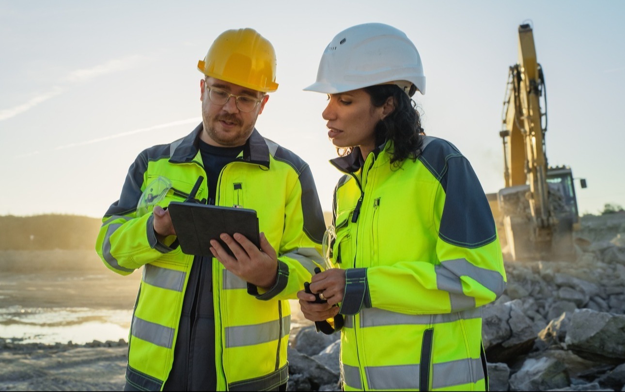 Male and female construction workers, on site with excavator in background, looking at tablet-1-3-1